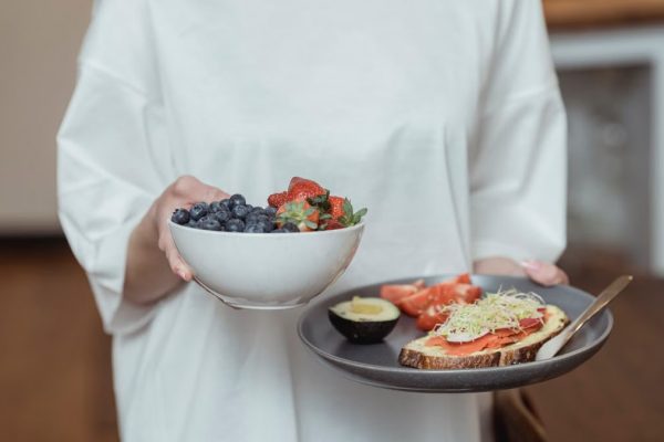 Person holding a bowl of fresh berries and a plate with avocado toast, displaying a healthy breakfast spread.