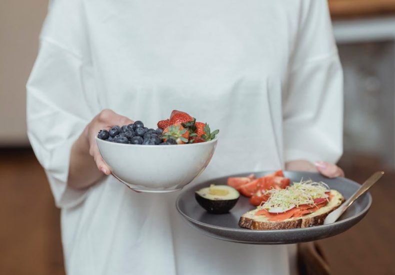 Person holding a bowl of fresh berries and a plate with avocado toast, displaying a healthy breakfast spread.
